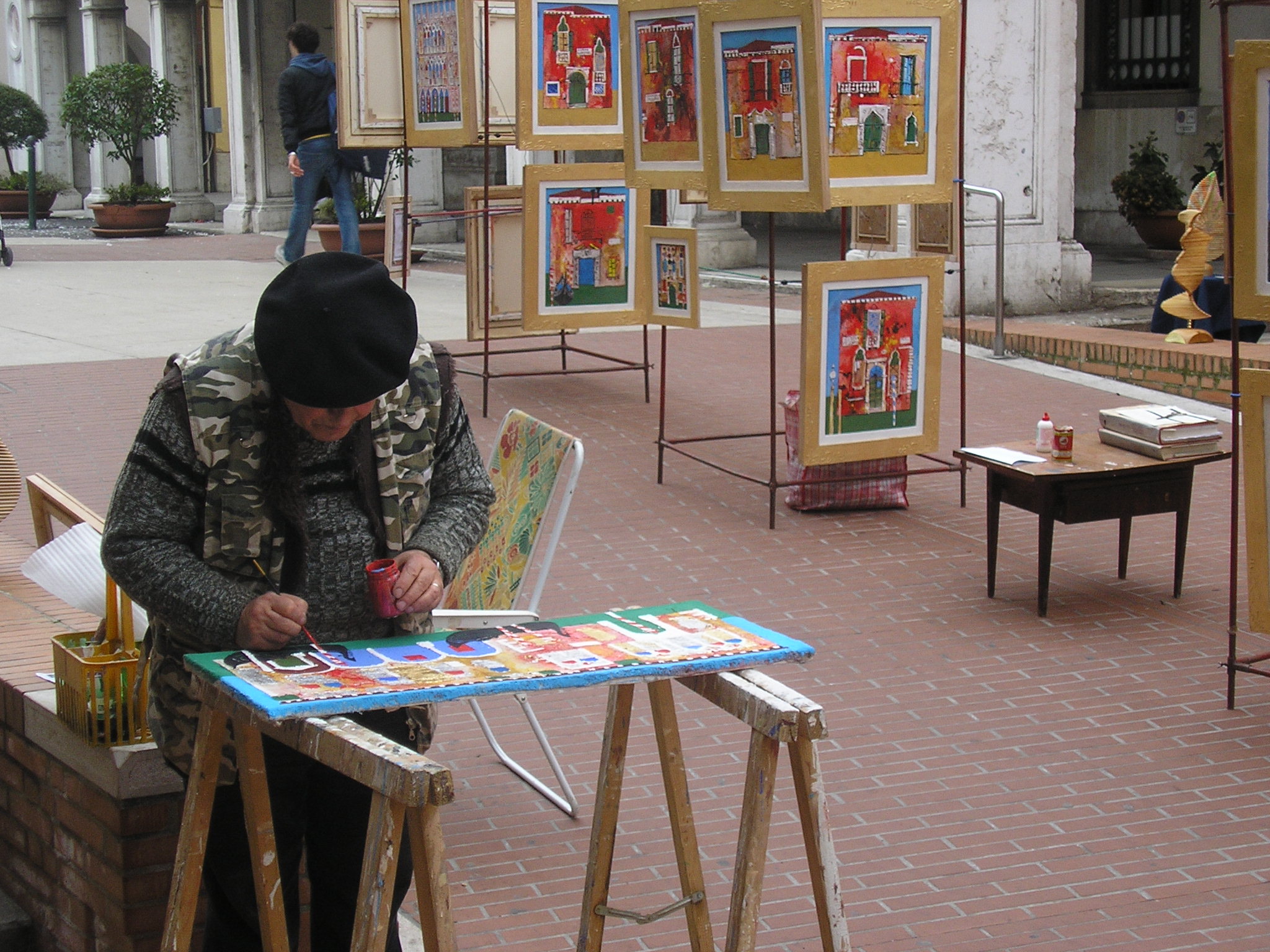 In primo piano pittore lavora a un quadro. Sullo sfondo alcuni quadri su cavalletti, esposti in strada
