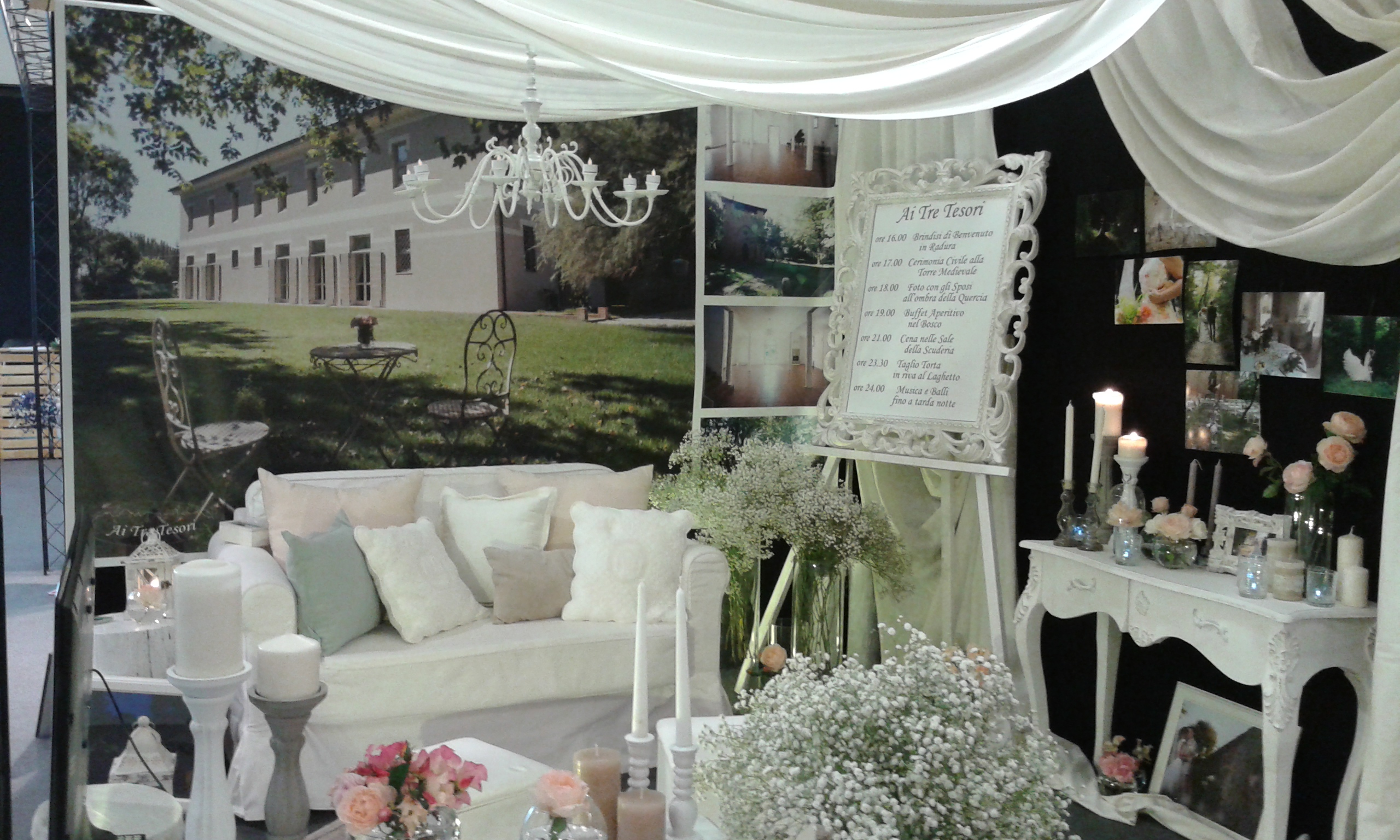 Stand di una fiera con divano e mobili bianchi, candele, fiori, foto e sullo sfondo l'immagine di una villa con giardino