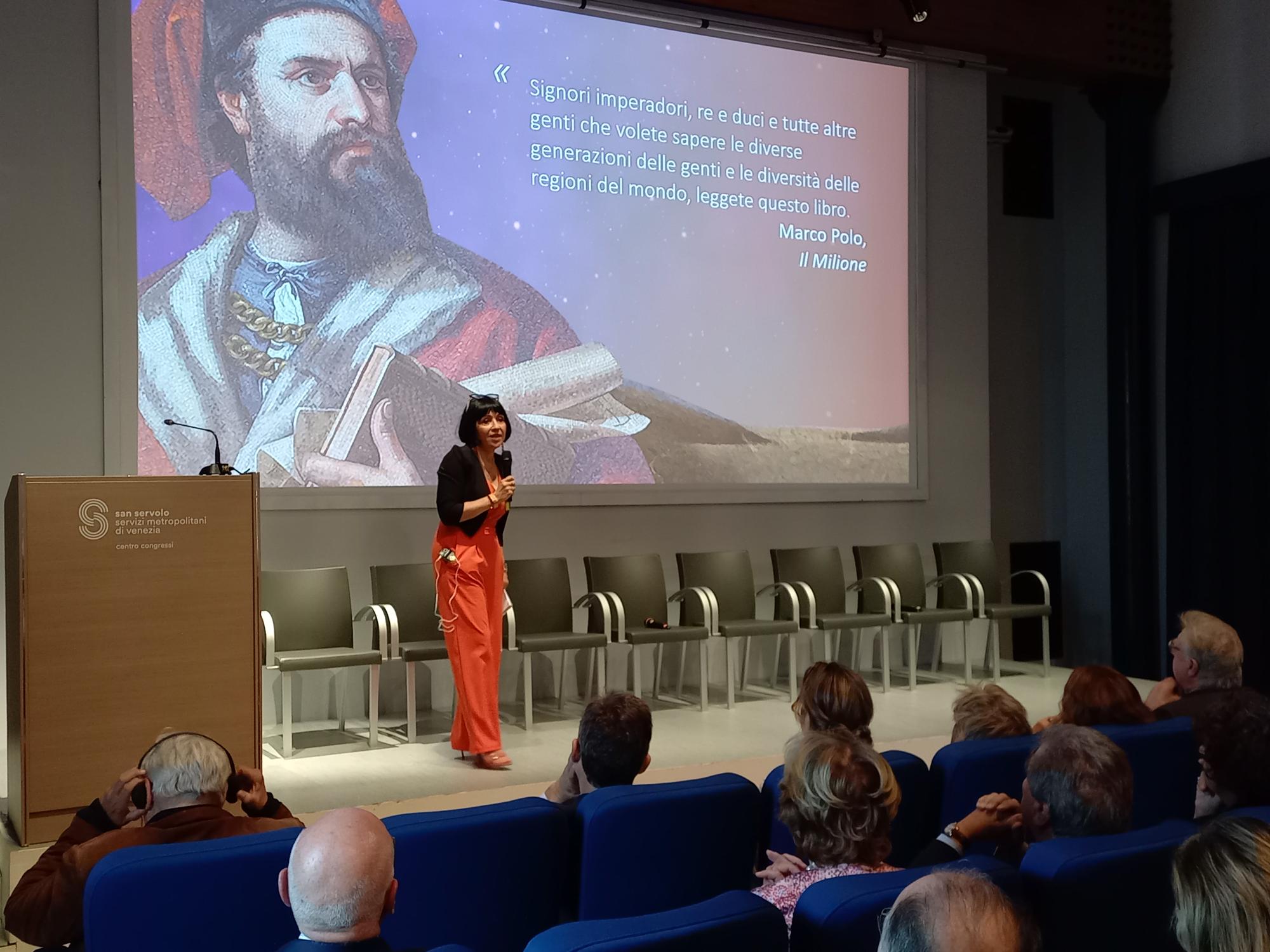 Donna parla con microfono al pubblico di un convegno. Dietro di lei, proiettata una slide con effigie di Marco Polo