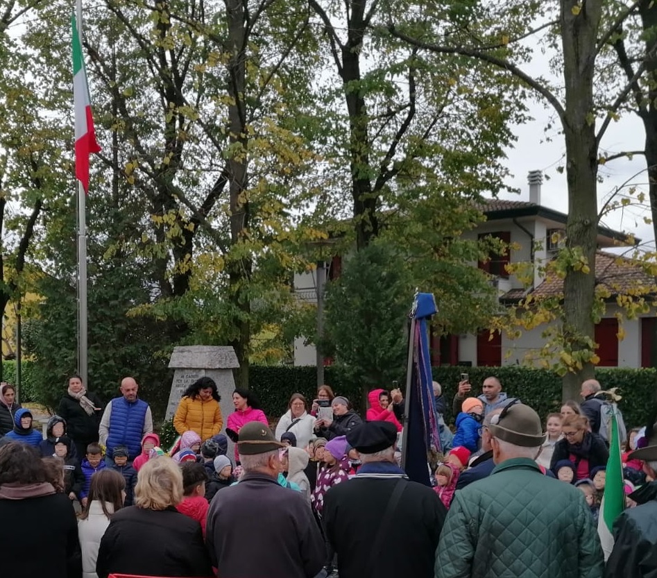 Folto gruppo di adulti e bambini. A sx Tricolore issato su pennone. In basso alcuni anziani con cappello da alpino