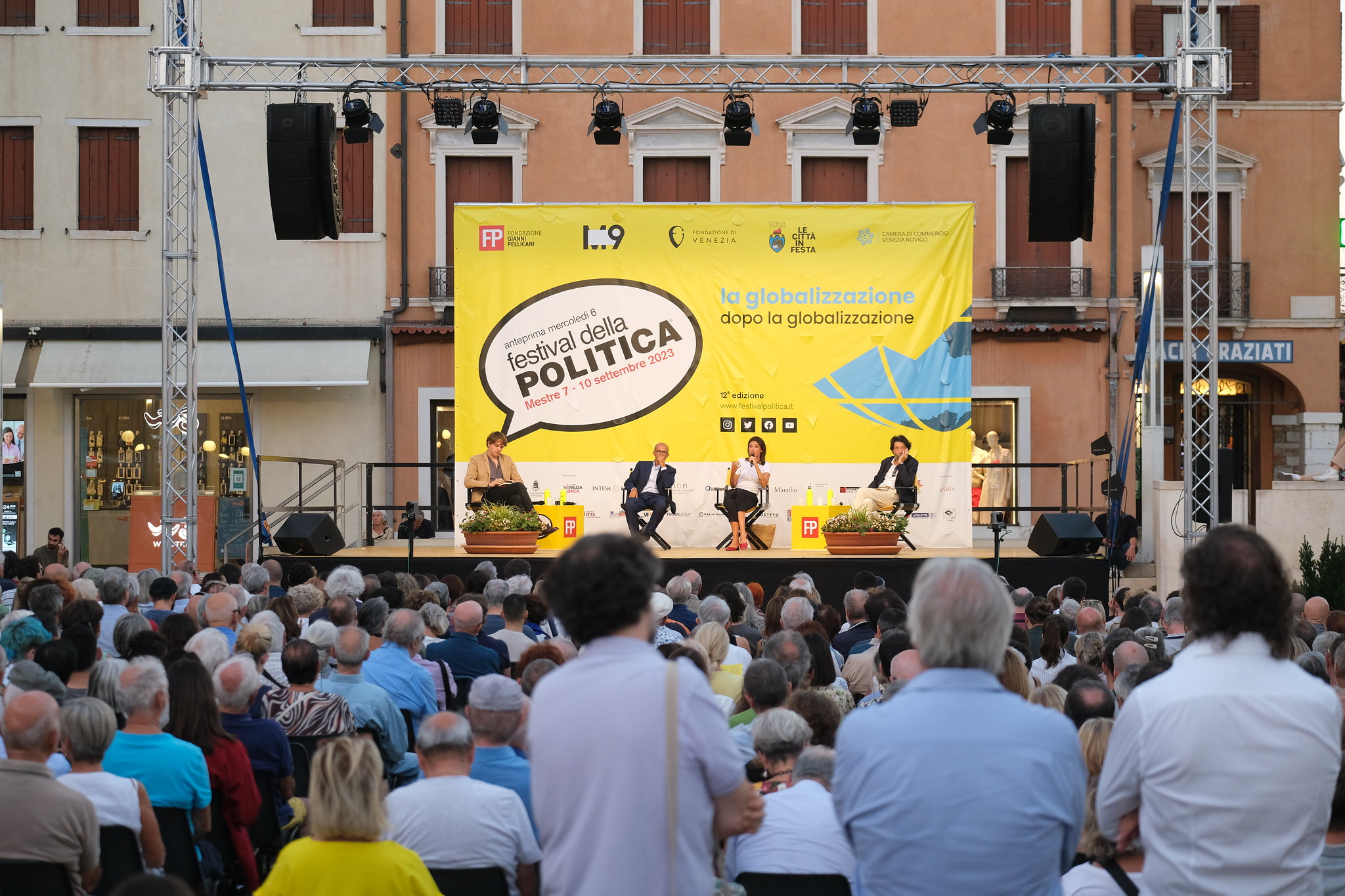 Tre uomini e una donna seduti su un palco, visti di fronte, parlano a un folto pubblico, inquadrato di spalle