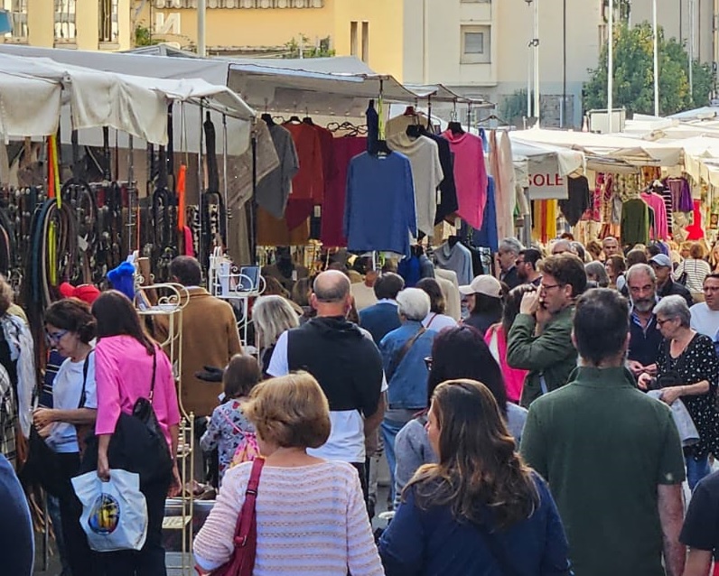 Folla di persone davanti ai banchi di un mercato all'aperto in una via cittadina