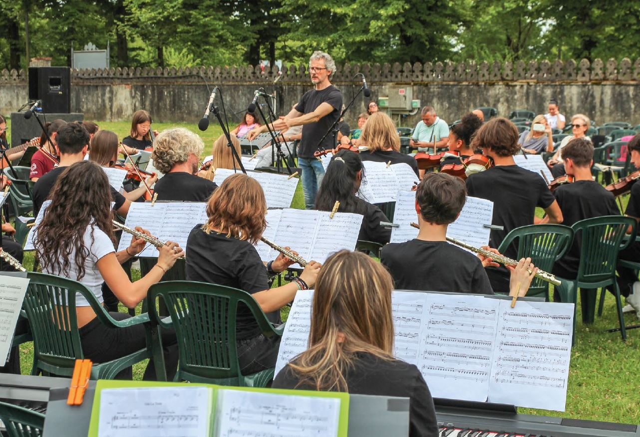 Orchestra di giovani, visti di spalle, suona all'aperto agli ordini di un direttore, in piedi, con gli occhiali