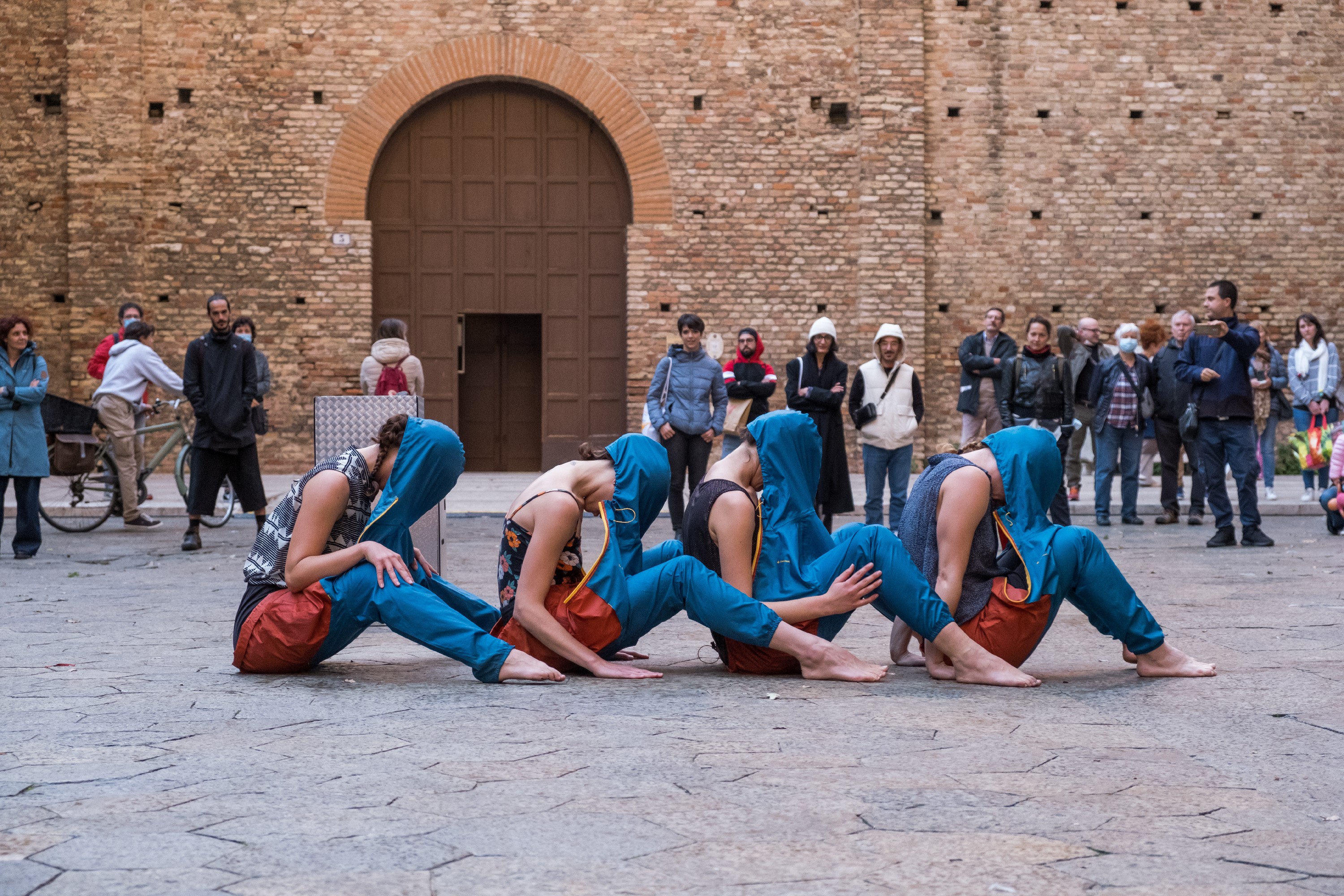 Quattro ballerine, in jeans e con un cappuccio a coprire il volto, sono sedute a terra in fila osservate dal pubblico