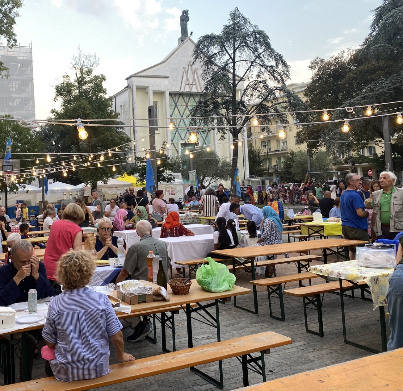 Persone sedute a tavoli da sagra sotto filari di lampadine. Sullo sfondo, una chiesa.