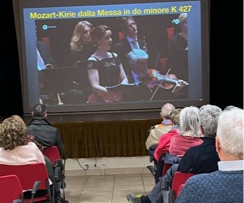 Pubblico visto di spalle osserva uno schermo su cui è proiettata l'immagine di alcuni musicisti