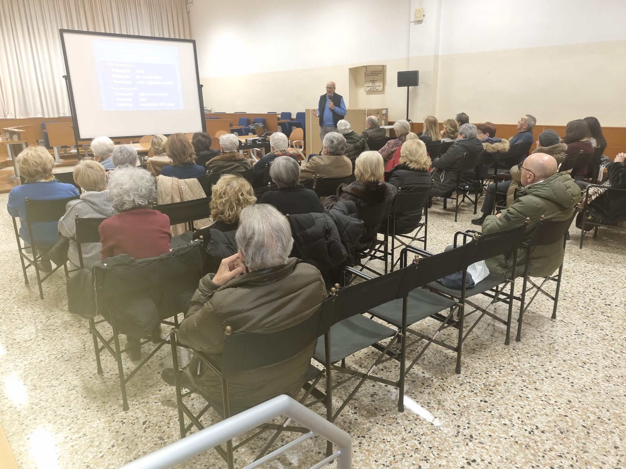 Pubblico di una conferenza inquadrato da dietro; davanti a uno schermo c'è il relatore che parla