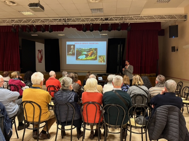 Pubblico seduto, visto di spalle, ascolta il relatore di una conferenza che parla in piedi al microfono