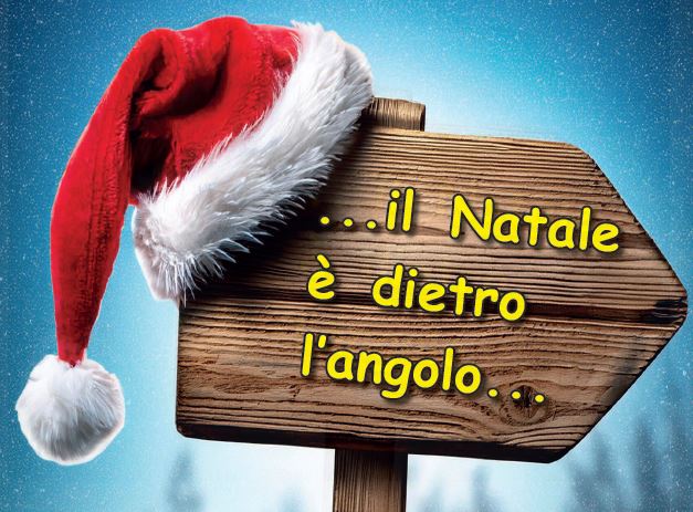 Cartello di legno con la scritta "...il Natale è dietro l'angolo" su cui è appeso un cappello da Babbo Natale