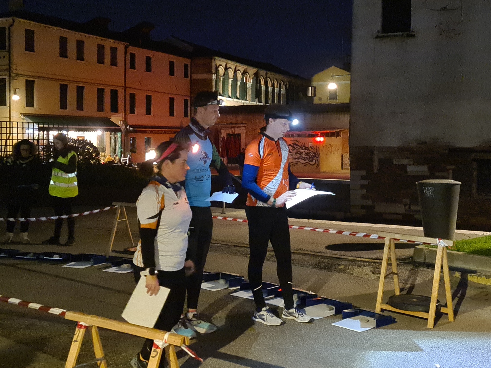 Due uomini e una donna alla partenza di una gara di orienteering, in abiti da sport e con in mano un foglio