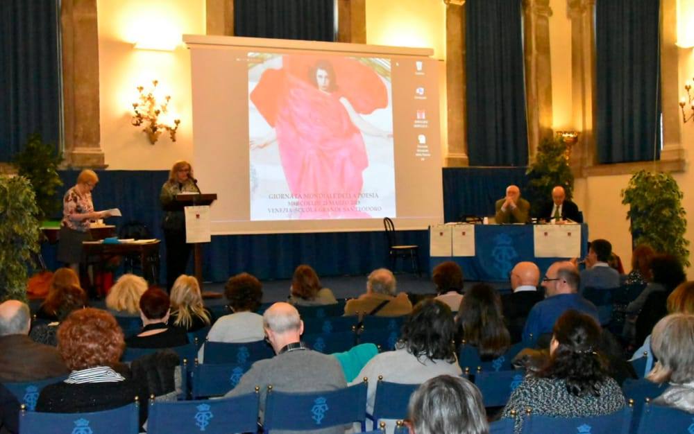 ubblico seduto ascolta un'oratrice sul palco, con dietro uno schermo che proietta una diapositiva