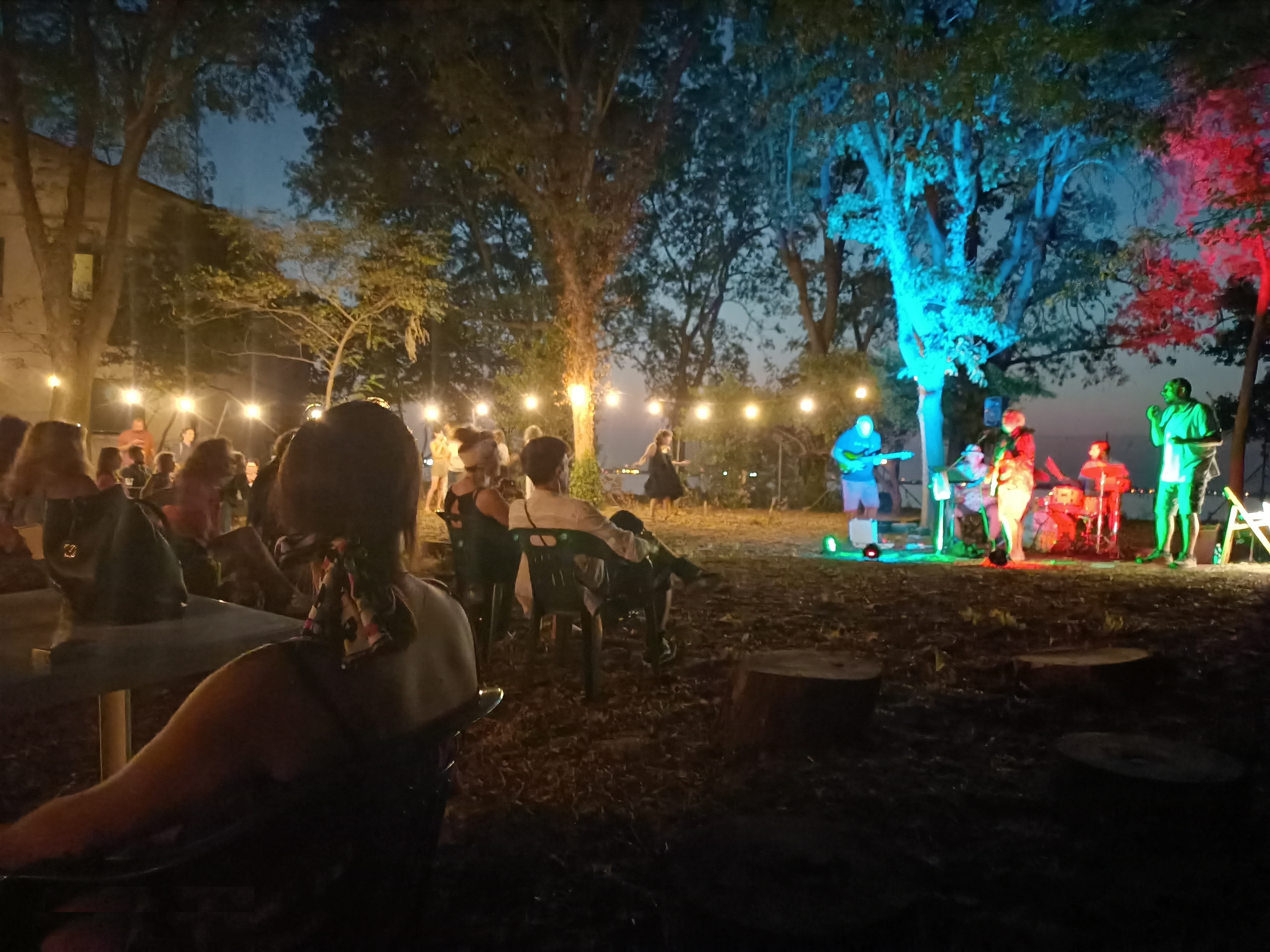 Gruppo musicale si esibisce di sera all'aperto in un parco di fronte a un pubblico seduto a tavolini