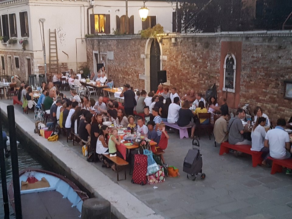 Lunghe tavolate gremite di persone allestite lungo una riva di Venezia