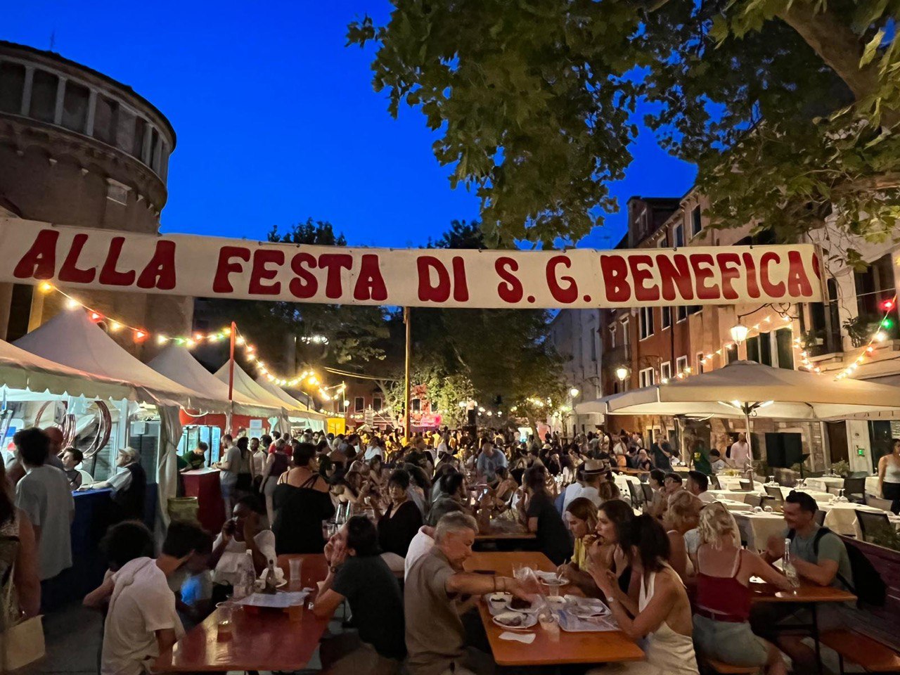 Una moltitudine di persone mangia seduta a tavoli allestiti in un campo veneziano, sotto striscione "Festa di S.G. Benefica"