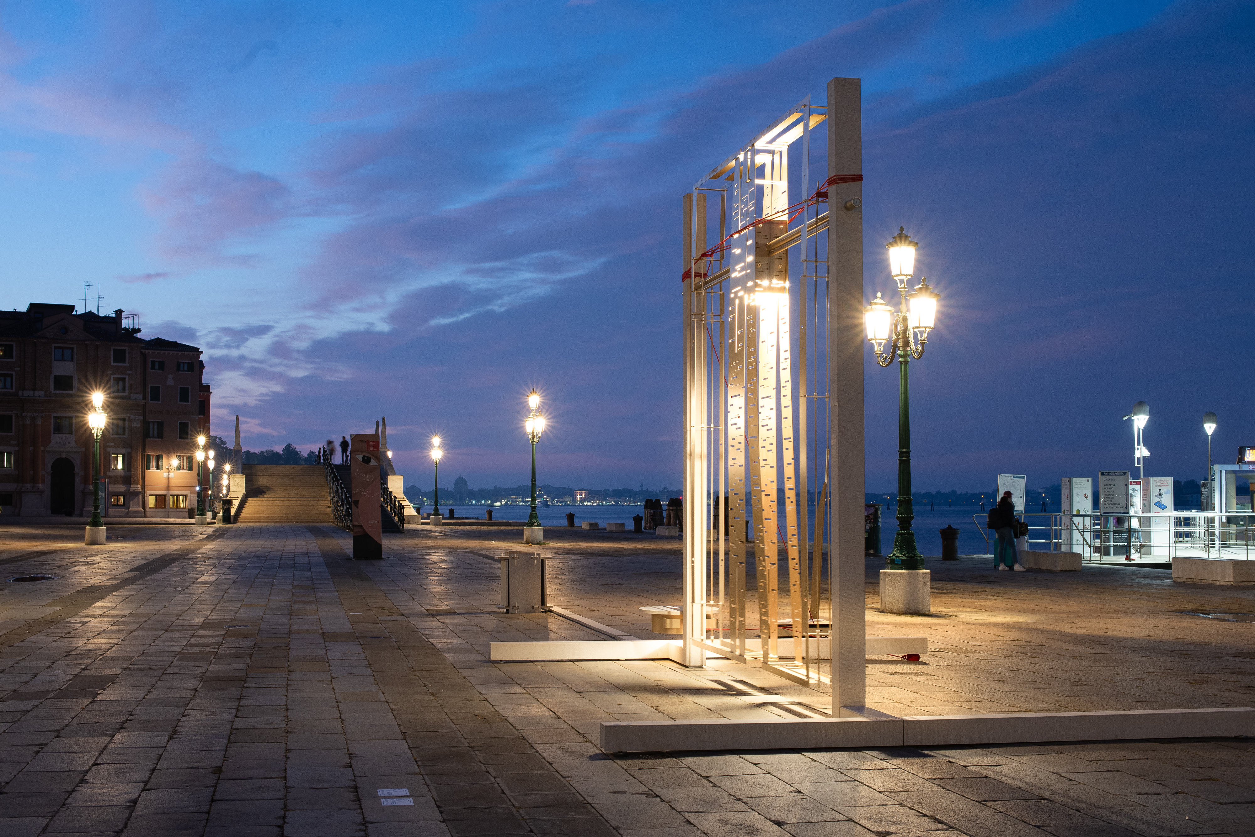 Paseaggio veneziano notturno: installazione artistica costituita da colonne e sbarre su una riva; sullo sfondo la laguna