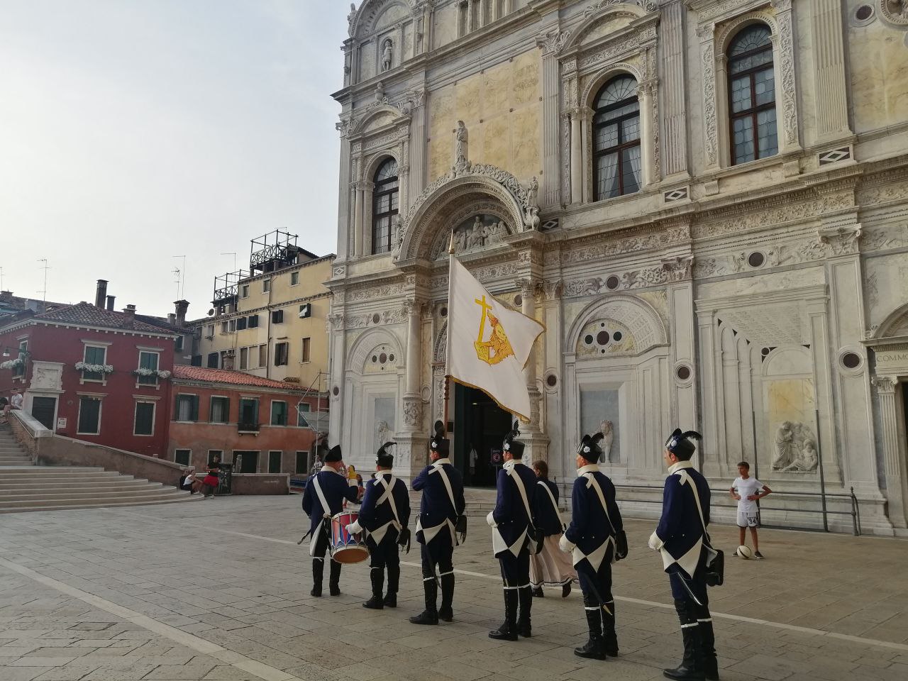 Sei uomini in abiti da soldati del '700 in fila in un campo veneziano, di fronte a una chiesa